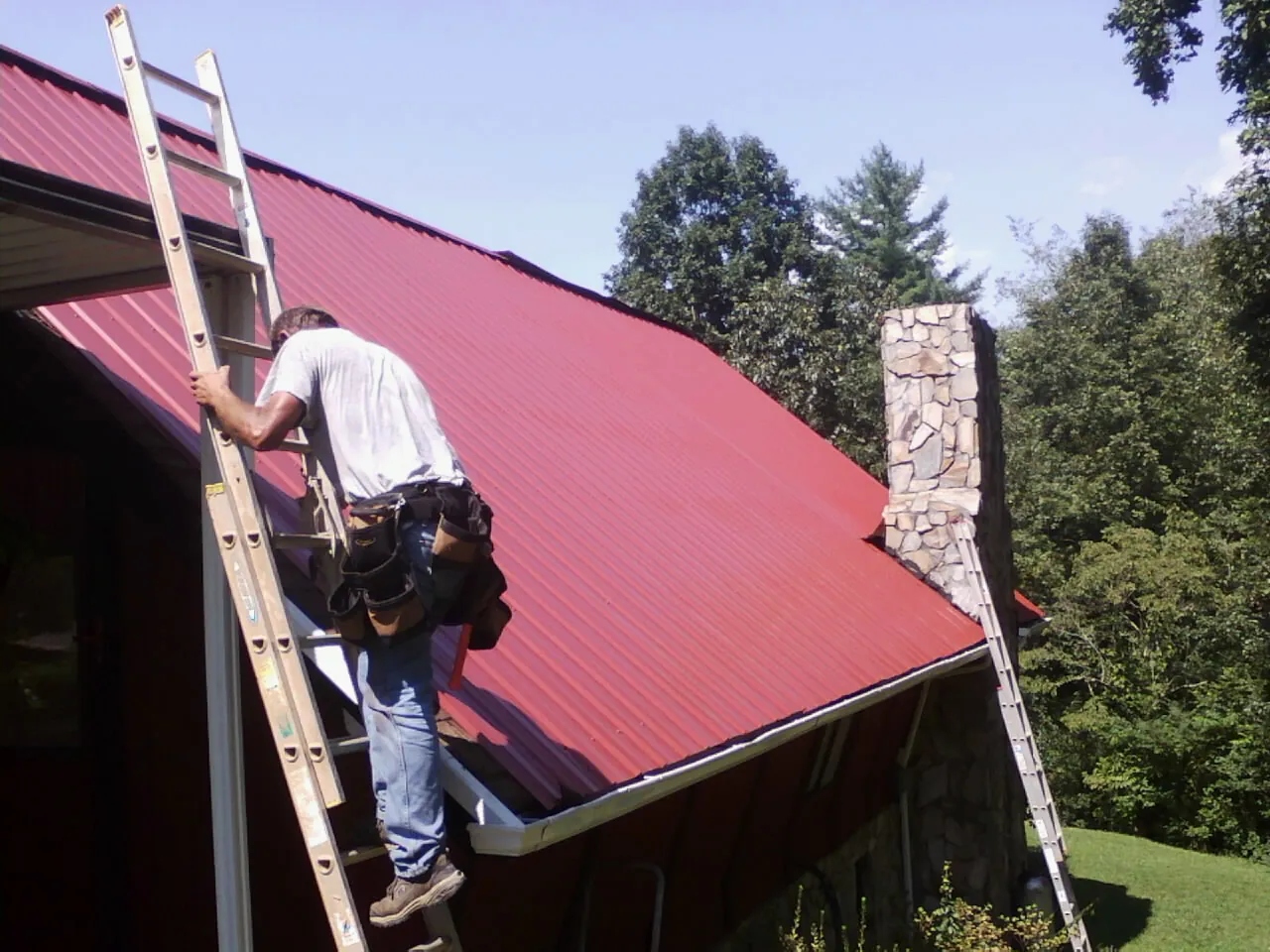 Roofer on a ladder beside a red metal roof and stone chimney — Soffit & Fascia Repair by Roofing Brothers in Palisades